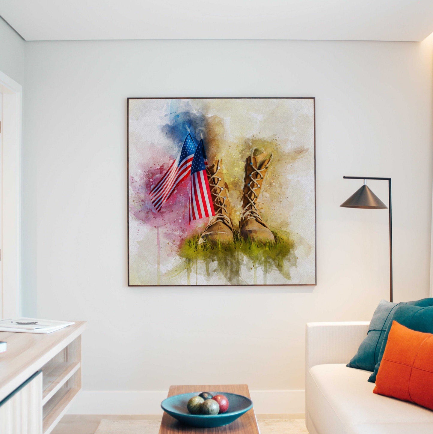 American Flag Wall Art: A framed watercolor print depicting two American flags and military boots in grass, hanging on a light-colored wall above a modern living room setting with a white couch, colorful pillows, a minimalist lamp, and a coffee table with a decorative bowl.