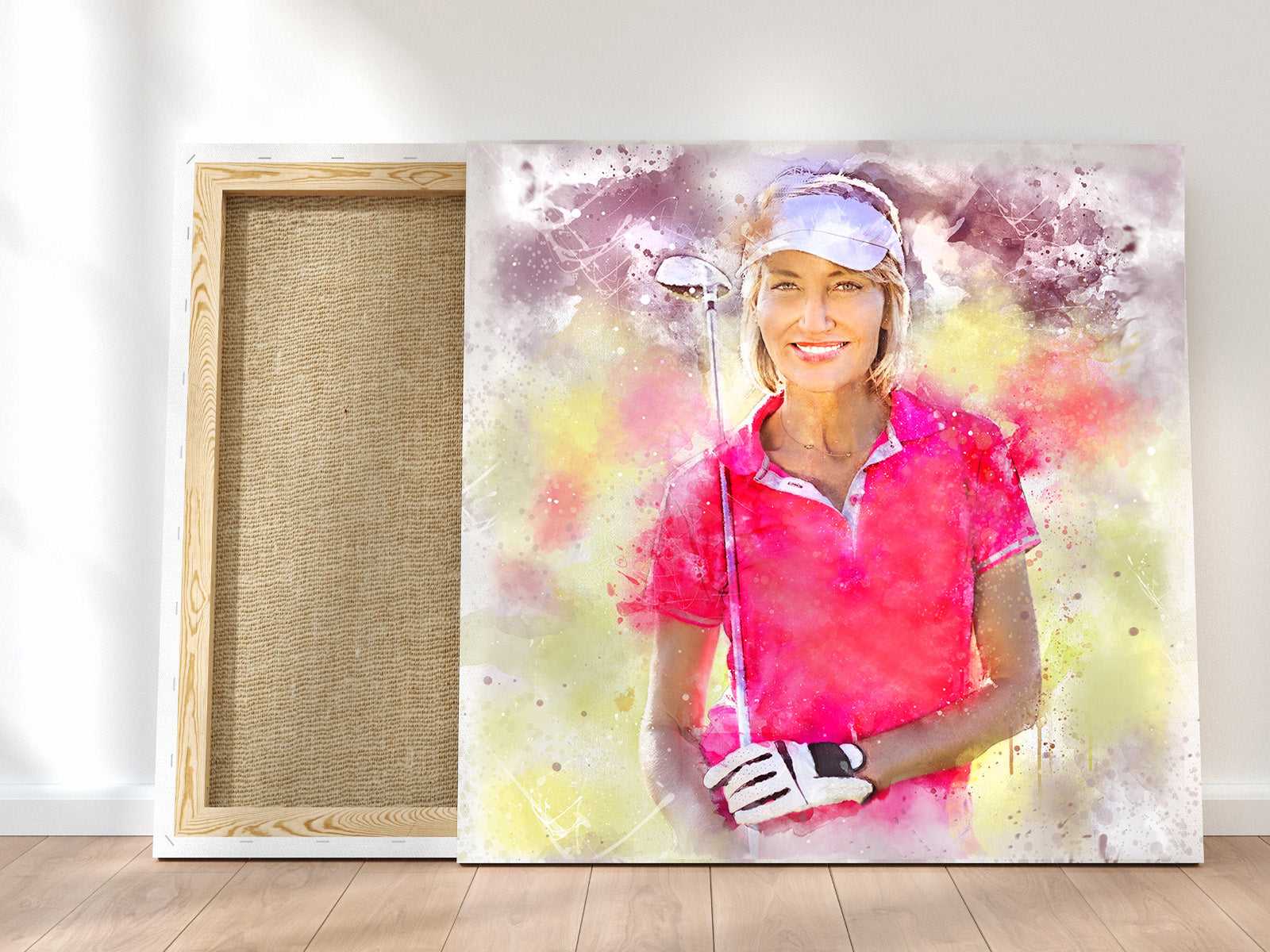 A smiling female golfer in a pink shirt and white visor, holding a club, is depicted in a vibrant watercolor golf art canvas leaning against a wall, with the back of another canvas visible.