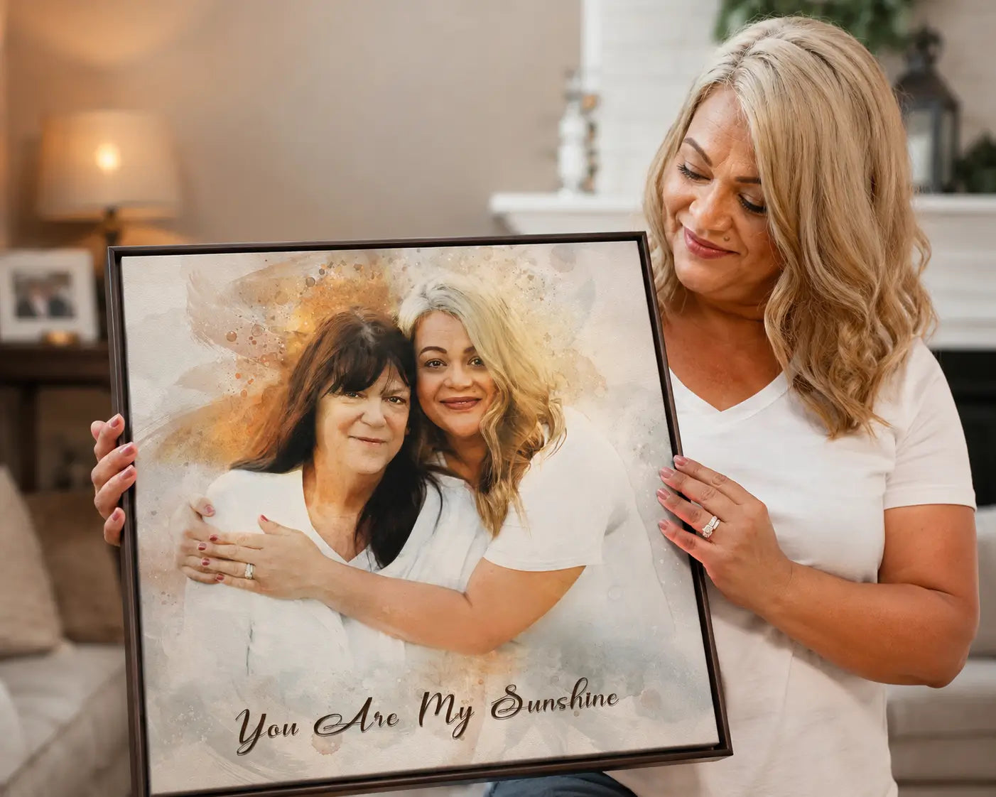 Woman holding a framed watercolor memorial portrait canvas featuring two women embracing with the text “You Are My Sunshine” in a cozy living room.